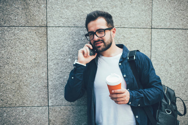 Pensive caucasian hipster guy in eyewear havin smartphone conversation standing near wall promotional background , handsome male student casual dressed making call in roaming on coffee break - Foto, Bild