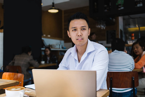 Adult pensive ethnic male in white shirt working remotely sitting at table with and mug of coffee working on laptop in cafe looking away  - Foto, Bild