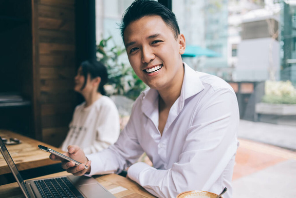 Young Asian male with smartphone in white shirt sitting at wooden desk with laptop and smiling while looking at camera in cafe against windows - Foto, Bild