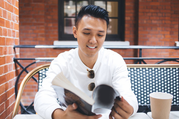Young smiling Asian male in white shirt holding book in hands and reading index while sitting at table with cup of coffee in cafe with brick walls - Фото, изображение