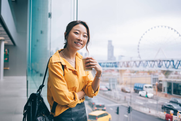Hermosa mujer asiática vistiendo camisa amarilla y falda de cuero negro dentada sonriendo y de pie junto a una ventana panorámica con vista a la rueda ferris sosteniendo café para ir y mirando a la cámara
 - Foto, imagen