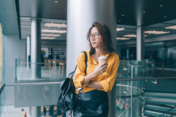Pensive Chinese hipster girl in classic glasses standing at public place and feeling pondering during free time indoors, attractive contemplative female in optical eyewear for provide eyes protection - Foto, imagen