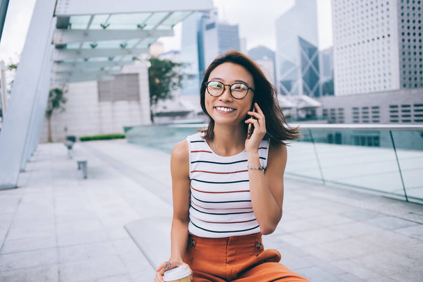 Half length portrait of joyful woman in optical glasses smiling at camera during international cell conversation using 4g internet in roaming, happy female tourist phoning via cellphone gadget - Zdjęcie, obraz