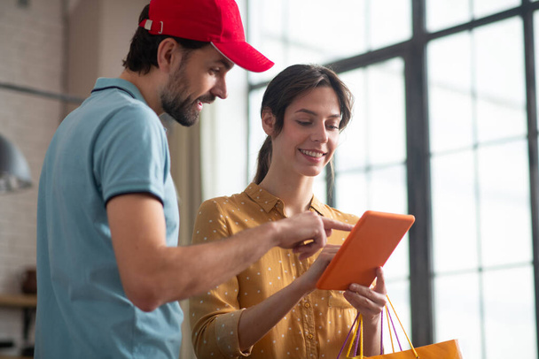 Deliver person in a red hat showing something on a tablet to a female customer - Photo, Image