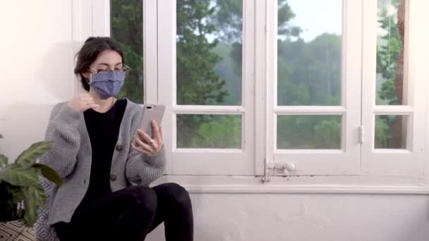Coronavirus. Woman in quarantine for coronavirus wearing protective mask, she's working from home and using mobile phone at sofa. Video call. Video conference with mobile phone. - Video, Çekim