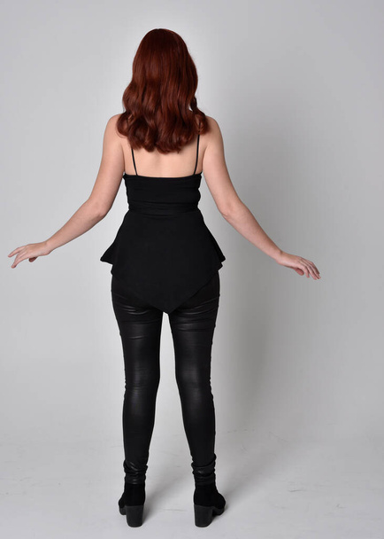  portrait of a pretty girl with red hair wearing black leather pants and top. Full length standing pose with back to the camera  isolated against a  grey studio background - Photo, image