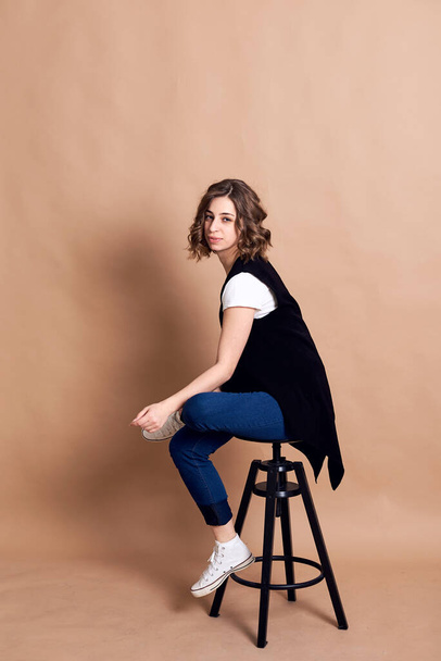 Young pretty brunette woman with curly hair, sitting on high black stool, posing, wearing white top, black vest and blue jeans. A portrait of Caucasian woman on beige background - 写真・画像