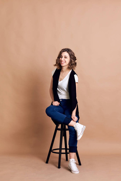 Young pretty brunette woman with curly hair, sitting on high black stool, posing, wearing white top, black vest and blue jeans. A portrait of Caucasian woman on beige background - Foto, Imagem
