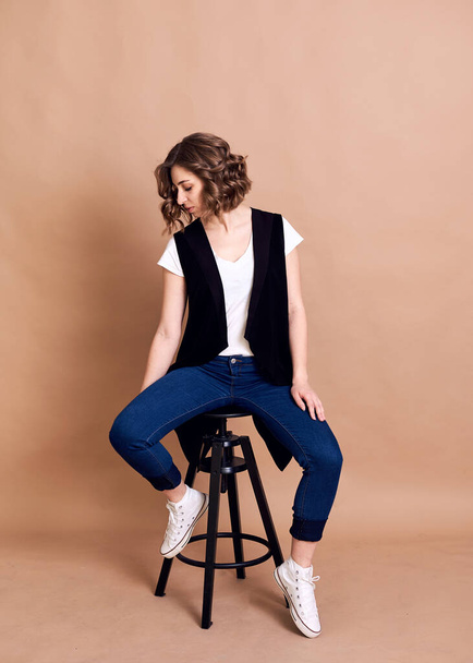 Young pretty brunette woman with curly hair, sitting on high black stool, posing, wearing white top, black vest and blue jeans. A portrait of Caucasian woman on beige background - Foto, afbeelding