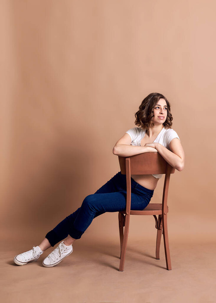 Young pretty brunette woman with curly hair, sitting on wooden brown stool, posing, wearing white top, black vest and blue jeans. A portrait of Caucasian woman on beige background - Photo, Image
