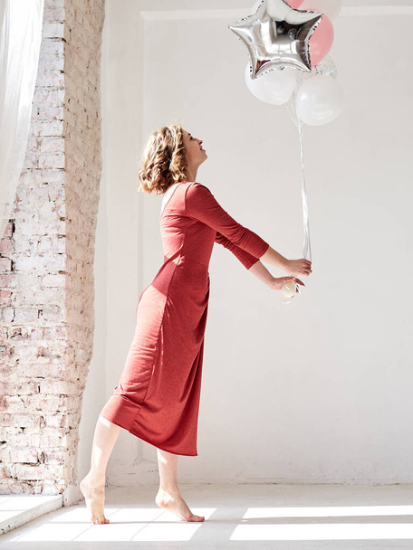 A full-length portrait of young pretty brunette woman with short curly hair, wearing red dress, doing dancing posing, holding pink and silver balloons, isolated on white background near big window - Foto, immagini