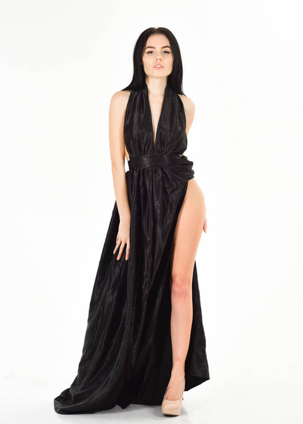 Woman in elegant black long evening dress with decollete, white background. Attractive girl wears expensive fashionable evening dress with erotic slit. Lady, sexy girl in dress. Fashion dress concept - Foto, imagen