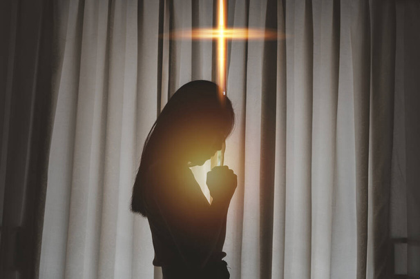 Silhouette φωτογραφία, Ασιάτισσα νεαρή γυναίκα, ενώ προσεύχεται λατρεία για τη χριστιανική θρησκεία στο δωμάτιο με το πορτοκαλί φως σταυρό, χριστιανική θρησκεία έννοια - Φωτογραφία, εικόνα