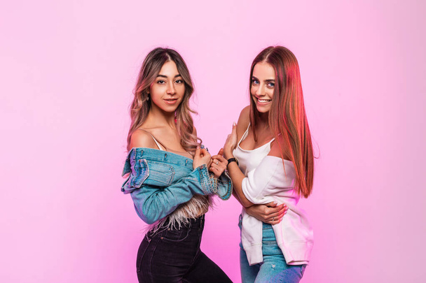 Two pretty happy women with positive smiles in trendy youth jeans clothes posing and smiling on background glamorous pink wall in the room. Pair cute joyful girlfriends in denim casual wear indoors. - Foto, Bild