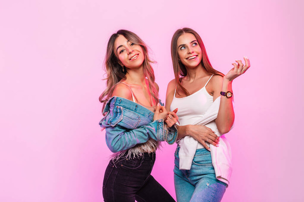 Two cute happy women with positive smiles in trendy youth jeans clothes posing and smiling on background glamorous pink wall in the room. Pair pretty joyful girlfriends in denim wear indoors. - Foto, Bild