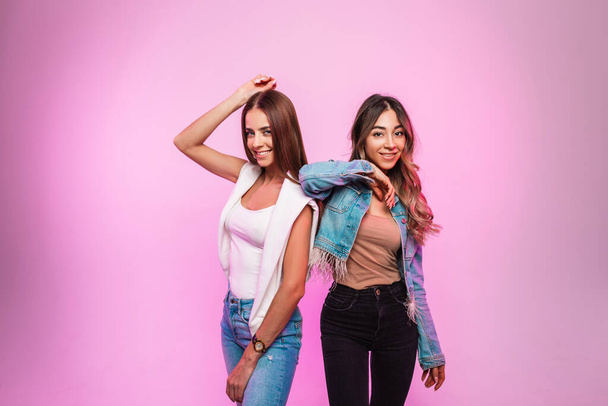 Grappige leuke positieve vriendinnen in trendy jeans kleding poseren in de buurt van fel roze muur in de studio. Gelukkige vrouwen in modieuze casual denim kleding staan en vrolijk glimlachen in de kamer. Voorjaarsstijl. - Foto, afbeelding