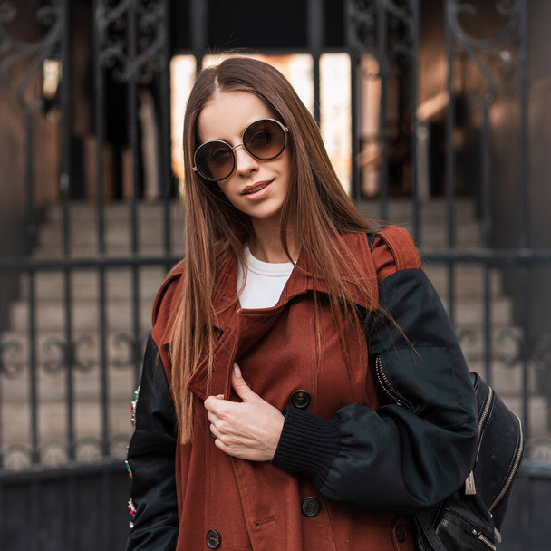 Elegant pretty young hipster woman in stylish sunglasses in fashionable brown trench coat with trendy leather black backpack walks on city near the iron gate. Urban girl outdoors. Spring youth style. - Foto, Imagem