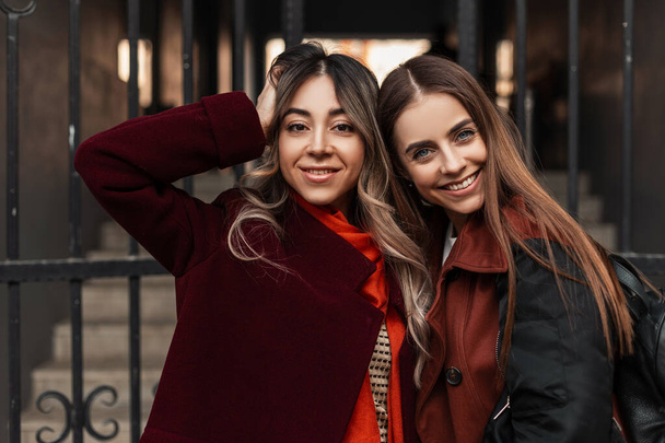 Portrait happy female friends with cute smile in fashionable spring clothes smiles near iron vintage gate in city. Cheerful pretty attractive girlfriends enjoying walk on street. Stylish urban ladies. - Photo, Image