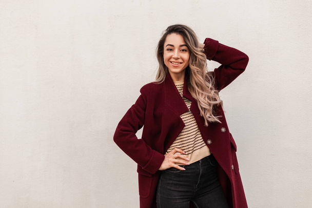 Joyful pretty young woman fashion model with long hair in stylish spring burgundy coat posing near vintage wall. Fashionable attractive girl with beautiful smile posing outdoors. Youth seasonal style - Photo, Image