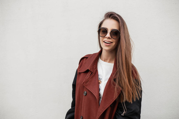 Pretty happy urban young woman with cute smile in fashionable spring youth clothes in stylish sunglasses posing near vintage wall in the city. American girl hipster smiling outdoors. Casual fashion. - Photo, Image