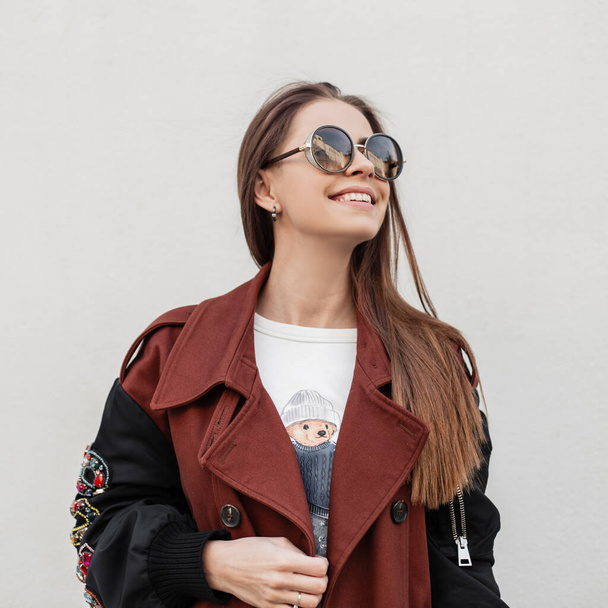 Pretty trendy joyful young woman hipster with cute smile in fashionable sunglasses in stylish spring trench coat poses near vintage wall in the city. Urban cheerful girl model enjoys a rest outdoors. - Foto, afbeelding