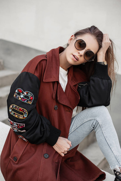 American young hipster woman in trendy sunglasses in stylish trench coat sits on steps and straightens hair. Beautiful girl in youth clothes is resting near vintage wall on the street in the city. - Photo, Image