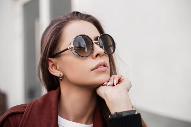 Portrét krásné mladé hipster žena v módní tmavé sluneční brýle ve stylovém hnědém plášti venku ve městě. Atraktivní dívka módní model s čistou pletí se sexy rty na ulici. Detailní záběr - Fotografie, Obrázek