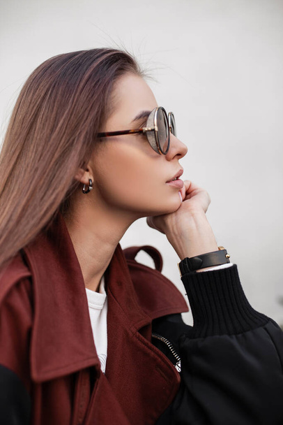 Modern urban young woman hipster with long hair with sexy lips in stylish sunglasses in fashionable brown spring trench coat near vintage wall in the city. Pretty elegant portrait female face.Close-up - Photo, Image