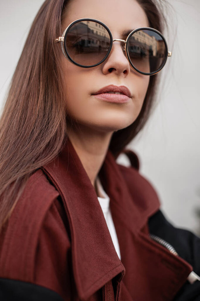 Piękny portret kobiety. Atrakcyjna młoda kobieta hipster z długimi włosami z seksownymi ustami w stylowych okularach przeciwsłonecznych w modnym brązowym wiosennym płaszczu odpoczywa w pobliżu zabytkowego budynku w mieście. - Zdjęcie, obraz