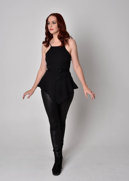 portrait of a pretty girl with red hair wearing black leather pants and top. Full length standing pose isolated against a  grey studio background - Photo, Image