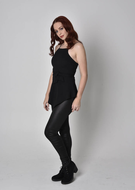 portrait of a pretty girl with red hair wearing black leather pants and top. Full length standing pose isolated against a  grey studio background - Zdjęcie, obraz