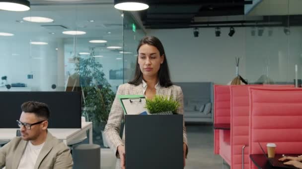 Smutná mladá žena opouští práci s krabicí věcí kráčející v otevřeném prostoru kanceláře - Záběry, video