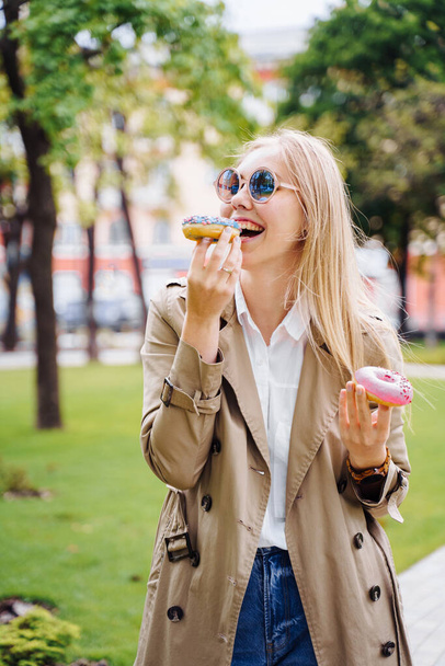 Junk food. Happy delighted woman in casual clothes eating donut with expression of big pleasure, temptation to bite doughnut, appetizing bakery. Sweet tooth,indulgence, gluttony, unhealthy lifestyle - Photo, Image