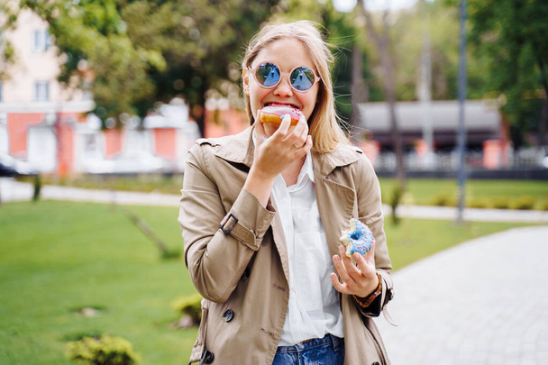 Непотрібна їжа. Щаслива приємна жінка в повсякденному одязі їсть пончик з виразом великого задоволення, спокусою вкусити пончик, апетитною пекарнею. Солодкий зуб, індульгенція, затишок, здоровий спосіб життя
 - Фото, зображення
