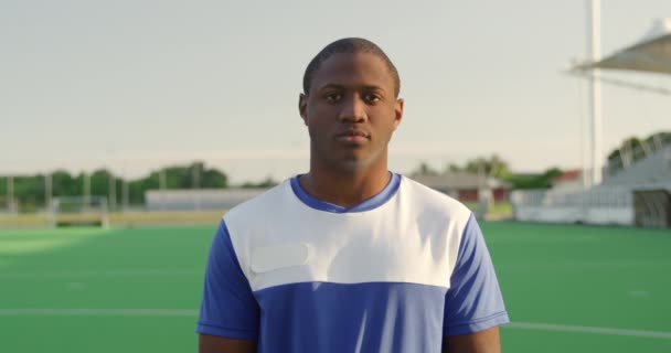 Portrait close up of an African American male field hockey player, wearing a blue team strip, standing on a hockey pitch looking to camera, smiling, on a sunny day, in slow motion - Video