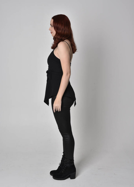  portrait of a pretty girl with red hair wearing black leather pants and top. Full length standing pose in side profile isolated against a  grey studio background - Foto, imagen