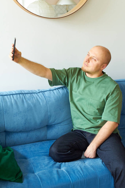 Man using smartphone and making selfie while sitting on blue sofa in living room - Фото, изображение