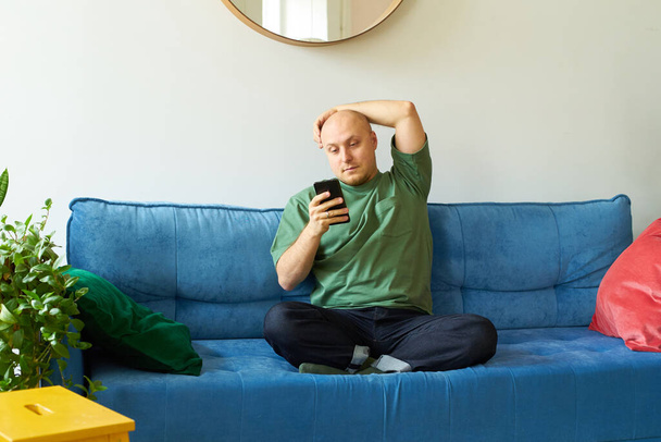 Man using smartphone while sitting on blue sofa in living room - Фото, изображение