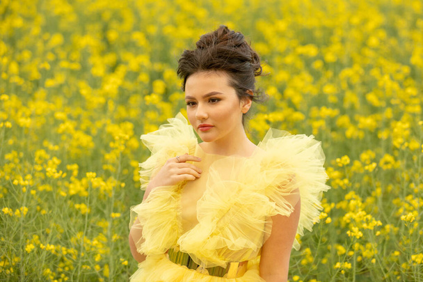 Portrait of romantic brunette teen girl wearing at yellow dress posing at field with yellow rapeseed flowers. . Happyness and summer time concept - Photo, Image
