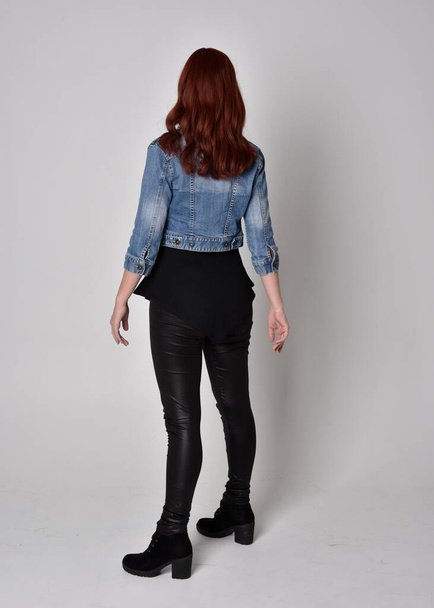 portrait of a pretty girl with red hair wearing black leather pants and denim jacket. Full length standing pose isolated against a  grey studio background - Photo, Image