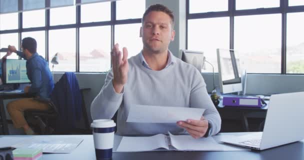 Front view of a Caucasian male business creative working in a casual modern office, talking, gesturing, holding a piece of paper during a video conference looking at camera, with colleagues in the background in slow motion - Кадры, видео