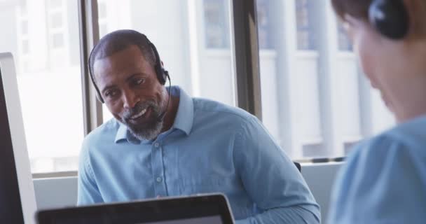 Front view of an African American male business creative working in a casual modern office, smiling and talking on a phone headset, with colleagues in the background in slow motion - Filmagem, Vídeo