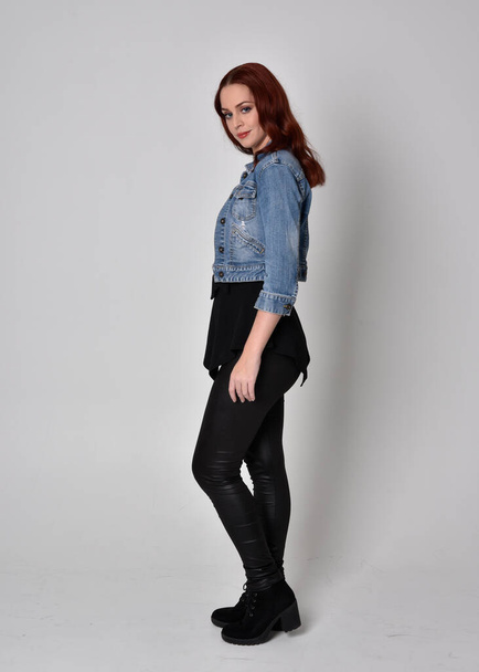 portrait of a pretty girl with red hair wearing black leather pants and denim jacket. Full length standing pose in side profile isolated against a  grey studio background - Zdjęcie, obraz