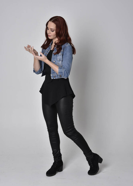 portrét hezké dívky s rudými vlasy v černých kožených kalhotách a džínové bundě. Plná délka stojící póza v bočním profilu izolované na šedém pozadí studia - Fotografie, Obrázek