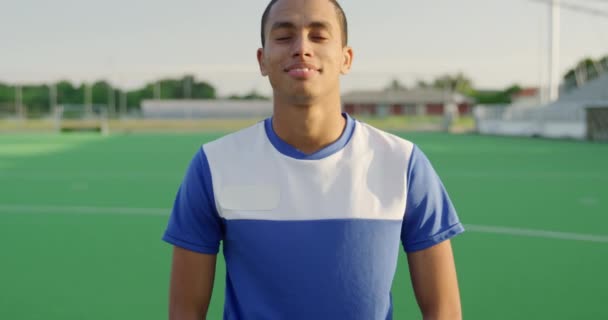 Portrait close up of a mixed race male field hockey player, wearing a blue team strip, standing on a hockey pitch looking to camera, smiling, on a sunny day, in slow motion - Video