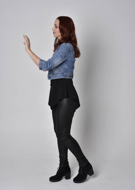 retrato de una chica bonita con el pelo rojo con pantalones de cuero negro y chaqueta de mezclilla. pose de pie de cuerpo entero en perfil lateral aislado sobre un fondo gris estudio
 - Foto, Imagen