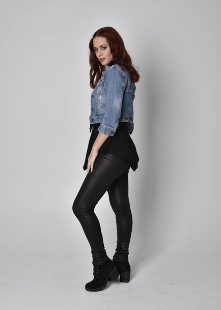 portrait of a pretty girl with red hair wearing black leather pants and denim jacket. Full length standing pose in side profile isolated against a  grey studio background - Photo, Image