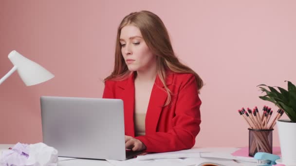 Μια συγκεντρωμένη νεαρή γυναίκα εργάζεται χρησιμοποιώντας τον φορητό υπολογιστή της κάθεται στο τραπέζι απομονωμένη σε ροζ φόντο - Πλάνα, βίντεο
