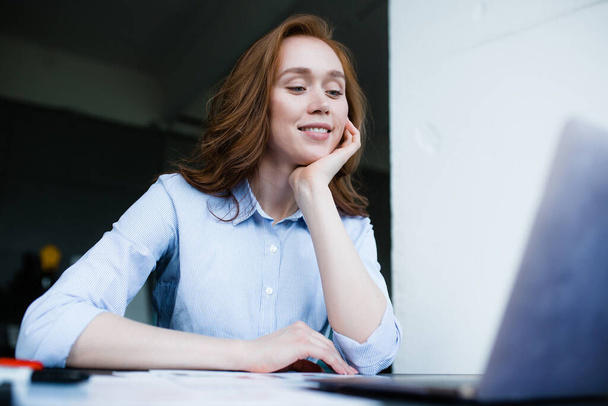 Redhead businesswoman put her chin on her hand and looks pretty at the laptop screen while doing her job. - Foto, Imagem