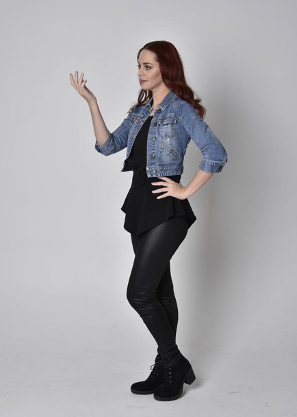  portrét hezké dívky s rudými vlasy v černých kožených kalhotách a džínové bundě. Plná délka stojící póza izolované na šedém pozadí studia - Fotografie, Obrázek